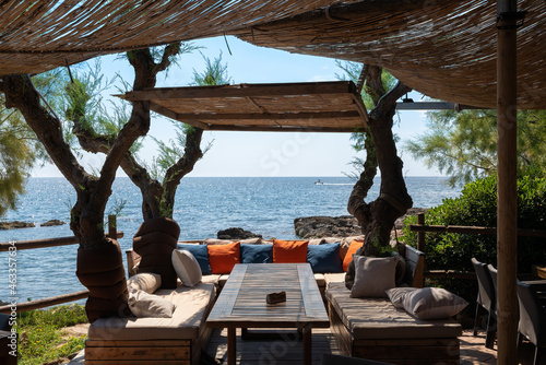 A table with comfortable benches to relax on the east coast of the Spanish island of Mallorca. There are many pillows on the cushions. In the background a boat is sailing across the water.