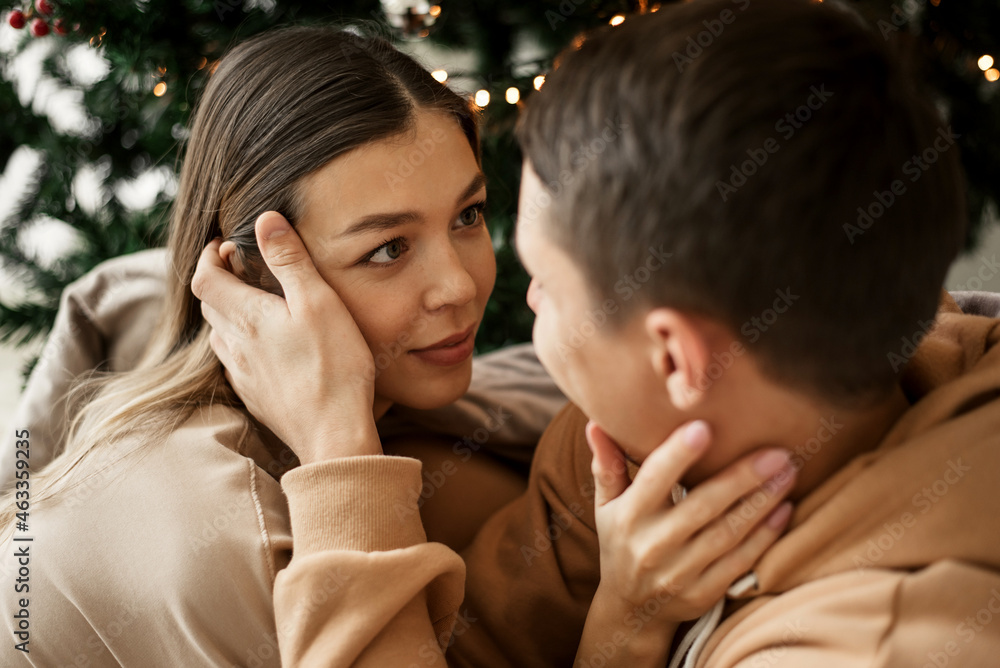 Cute young couple sitting near Christmas tree. Man and woman looking at each other Cute young couple sitting near Christmas tree. Man and woman looking at each other