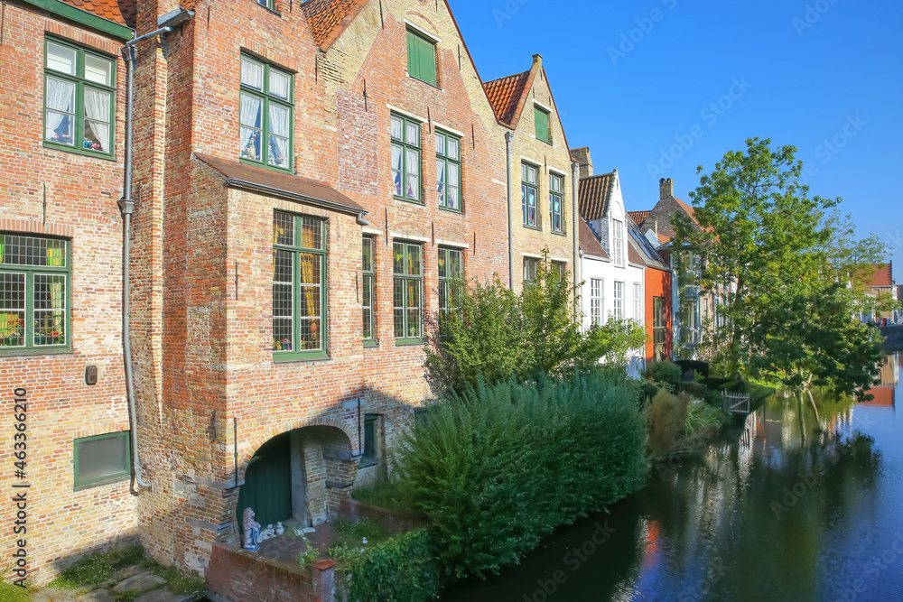 Brugge, Belgium - October 9. 2021: View along water canal with green trees and old residential brick house against clear blue sky