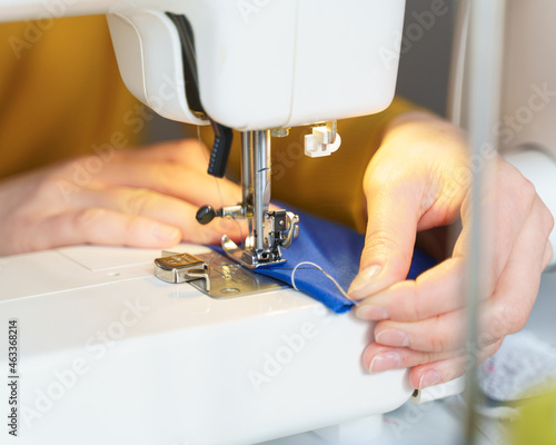 Close up shot of tailor or seamstress sewing or stitching blue navy fabric with professional machine while sitting and making new clothing item at her workplace in atelier or workshop, selective focus