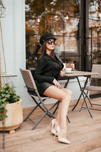 Brunette girl in a black cap in a leather jacket in a skirt and in beige high-heeled shoes on the street near the coffee shop in sunglasses © Alina Belych