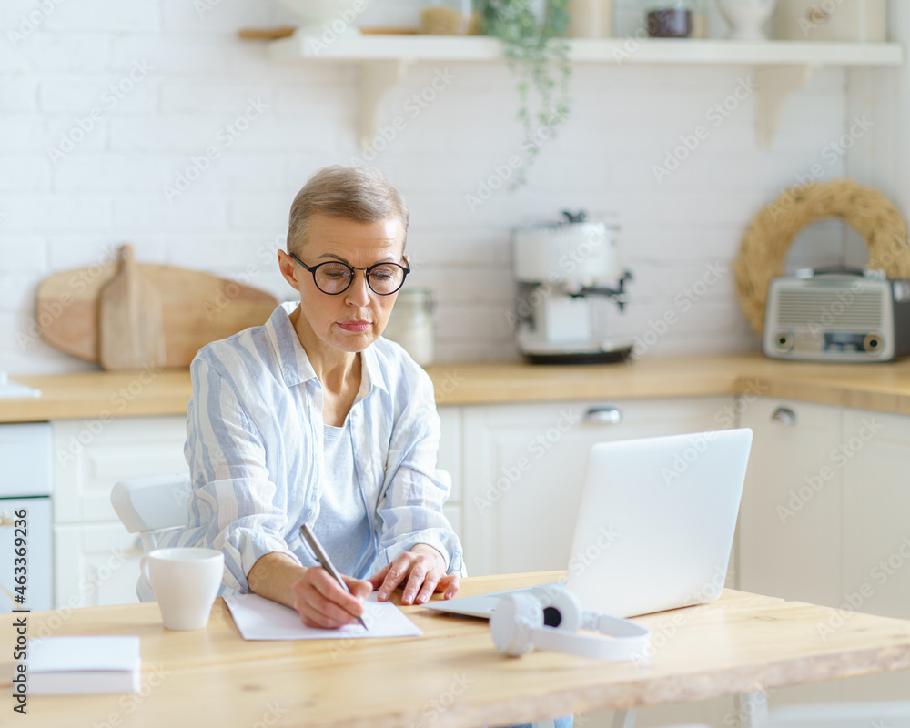 Modern focused mature woman writer wearing eyeglasses typing something on laptop, working online while sitting in kitchen at home. Senior people and technologies concept Modern focused mature woman writer wearing eyeglasses typing something on laptop, working online while sitting in kitchen at home. Senior people and technologies concept