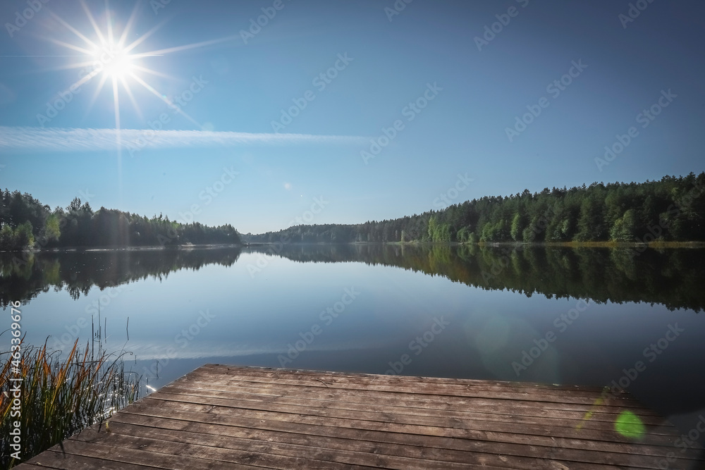 Summer landscape with green forest, lake water, clear sky with sun shining with rays and its reflection in river.