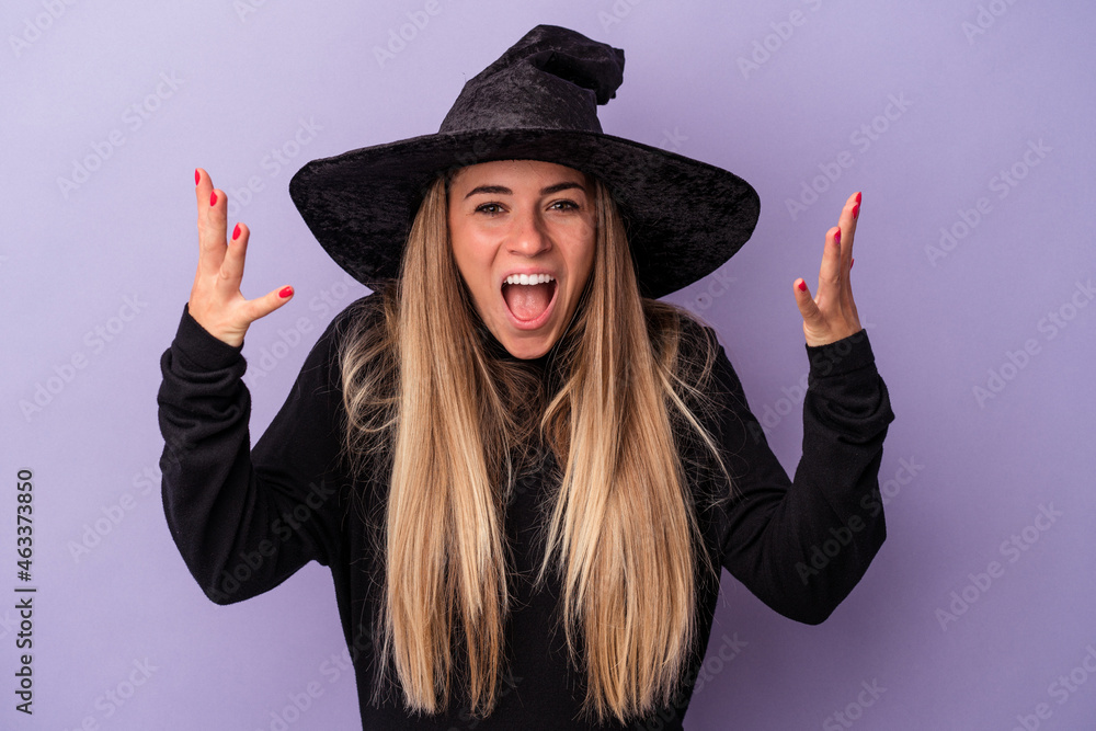 Young Russian woman disguised as a witch celebrating Halloween isolated on purple background receiving a pleasant surprise, excited and raising hands.