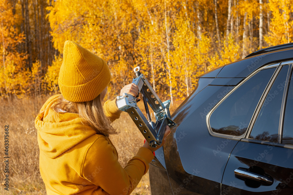 A woman in yellow clothes is trying to change a car wheel with a jack on a picnic in the woods on an autumn day. A woman in yellow clothes is trying to change a car wheel with a jack on a picnic in the woods on an autumn day.