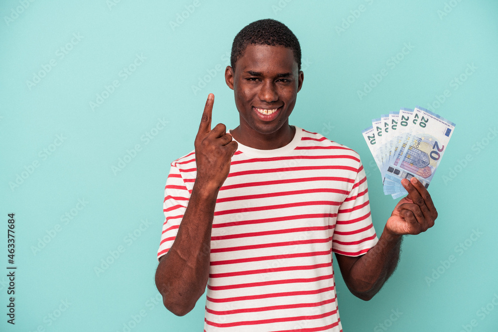 Young African American man holding a bank notes isolated on blue background showing number one with finger. Young African American man holding a bank notes isolated on blue background showing number one with finger.