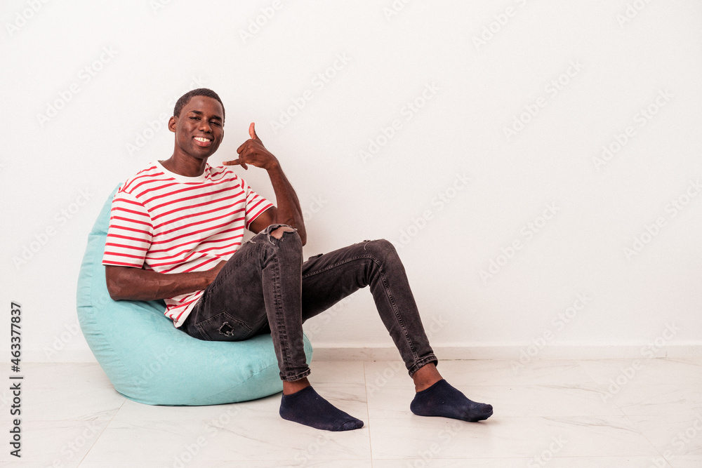 Fototapeta premium Young African American man sitting on a puff isolated on white background showing a mobile phone call gesture with fingers.