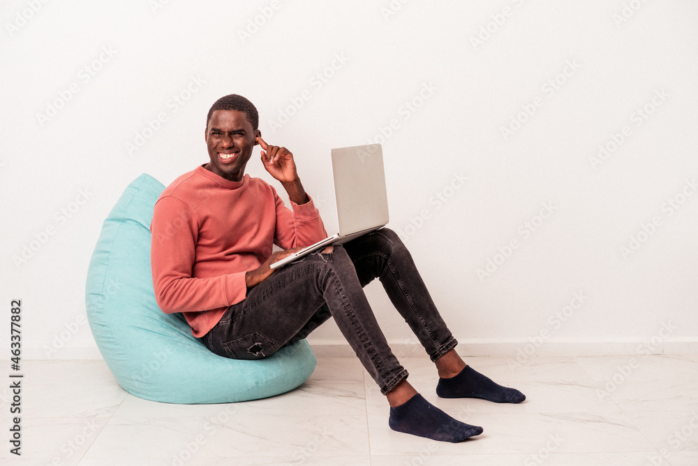 Young African American man sitting on a puff using laptop isolated on white background covering ears with hands.