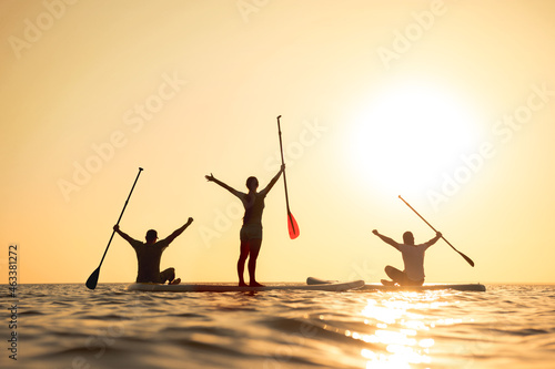 Happy surfers stands on sup boards with raised arms and looks at sunset