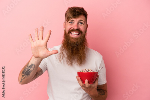 Young caucasian ginger man with long beard holding a bowl of cereales isolated on pink background smiling cheerful showing number five with fingers.