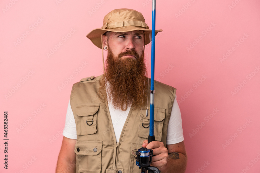 Young caucasian ginger fisherman with long beard holding a rod isolated on pink background confused, feels doubtful and unsure. Young caucasian ginger fisherman with long beard holding a rod isolated on pink background confused, feels doubtful and unsure.