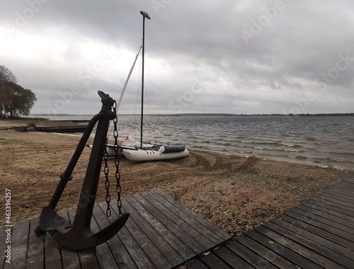 Big old rusty anchor on wooden pier on sandy shore of Volga river with inflatable boat and the river itself on the background