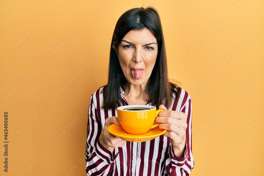 Young hispanic woman drinking a cup of coffee sticking tongue out happy with funny expression. Young hispanic woman drinking a cup of coffee sticking tongue out happy with funny expression.