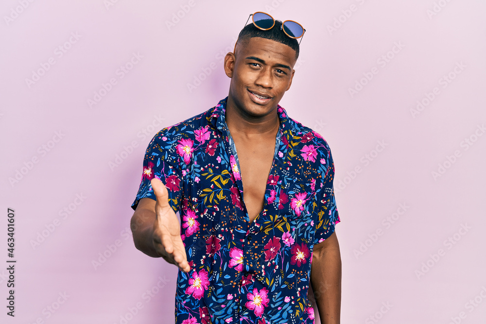 Fototapeta premium Young black man wearing hawaiian shirt and sunglasses smiling friendly offering handshake as greeting and welcoming. successful business.