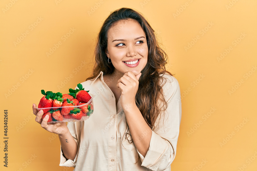 Young hispanic girl holding strawberries with hand on chin thinking about question, pensive expression. smiling and thoughtful face. doubt concept. Young hispanic girl holding strawberries with hand on chin thinking about question, pensive expression. smiling and thoughtful face. doubt concept.