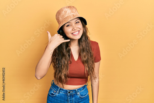 Young hispanic girl wearing casual clothes and hat smiling doing phone gesture with hand and fingers like talking on the telephone. communicating concepts.