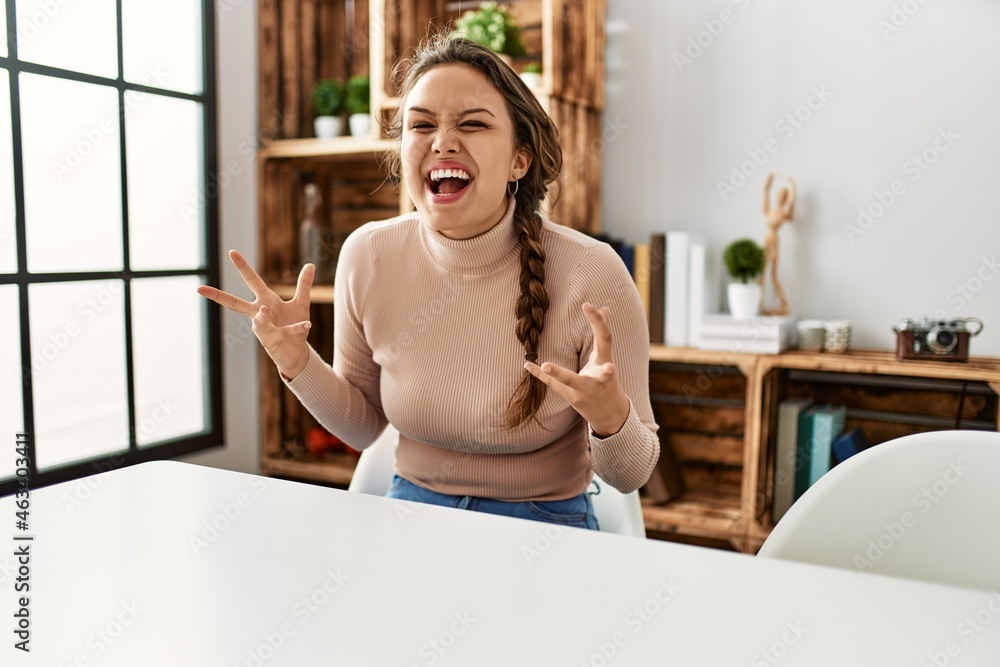 Young hispanic girl wearing casual clothes sitting on the table at home celebrating mad and crazy for success with arms raised and closed eyes screaming excited. winner concept Young hispanic girl wearing casual clothes sitting on the table at home celebrating mad and crazy for success with arms raised and closed eyes screaming excited. winner concept