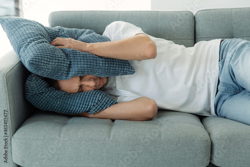 Fotografia Annoyed man tired of noisy sounds from neighbors try to sleep on couch covering head with pillows