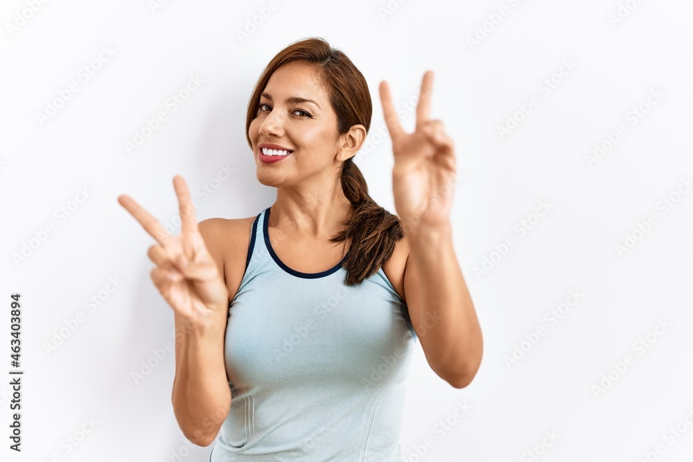 Young latin woman wearing sporty clothes over isolated background smiling looking to the camera showing fingers doing victory sign. number two. Young latin woman wearing sporty clothes over isolated background smiling looking to the camera showing fingers doing victory sign. number two.