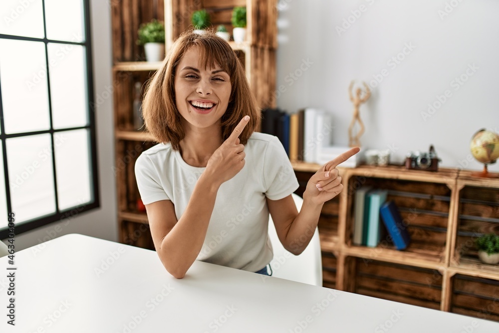 Young caucasian girl wearing casual clothes sitting on the table at home smiling and looking at the camera pointing with two hands and fingers to the side. Young caucasian girl wearing casual clothes sitting on the table at home smiling and looking at the camera pointing with two hands and fingers to the side.