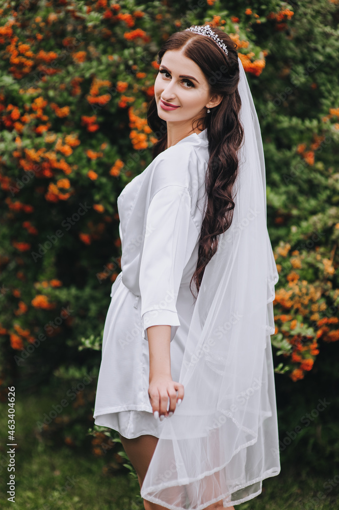 A beautiful, smiling bride model with curly hair in white lingerie, underwear walks, dances in nature, turning around in the park on green grass against a background of orange rowan. Wedding portrait.