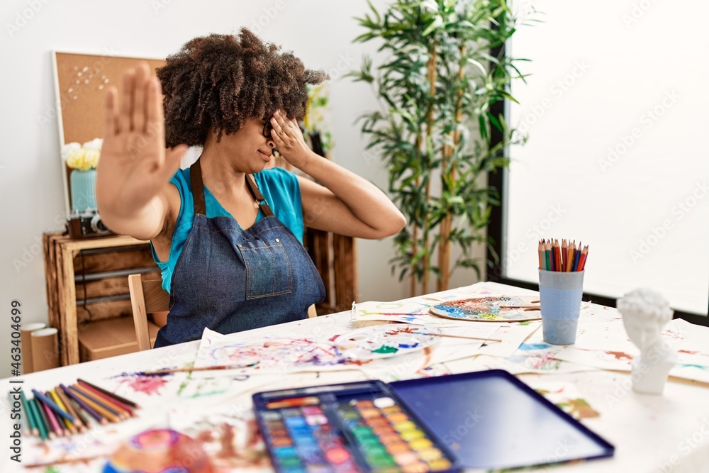 Beautiful african american woman with afro hair painting at art studio covering eyes with hands and doing stop gesture with sad and fear expression. embarrassed and negative concept.