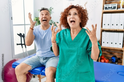 Physiotherapy woman working at pain recovery clinic with patient crazy and mad shouting and yelling with aggressive expression and arms raised. frustration concept.