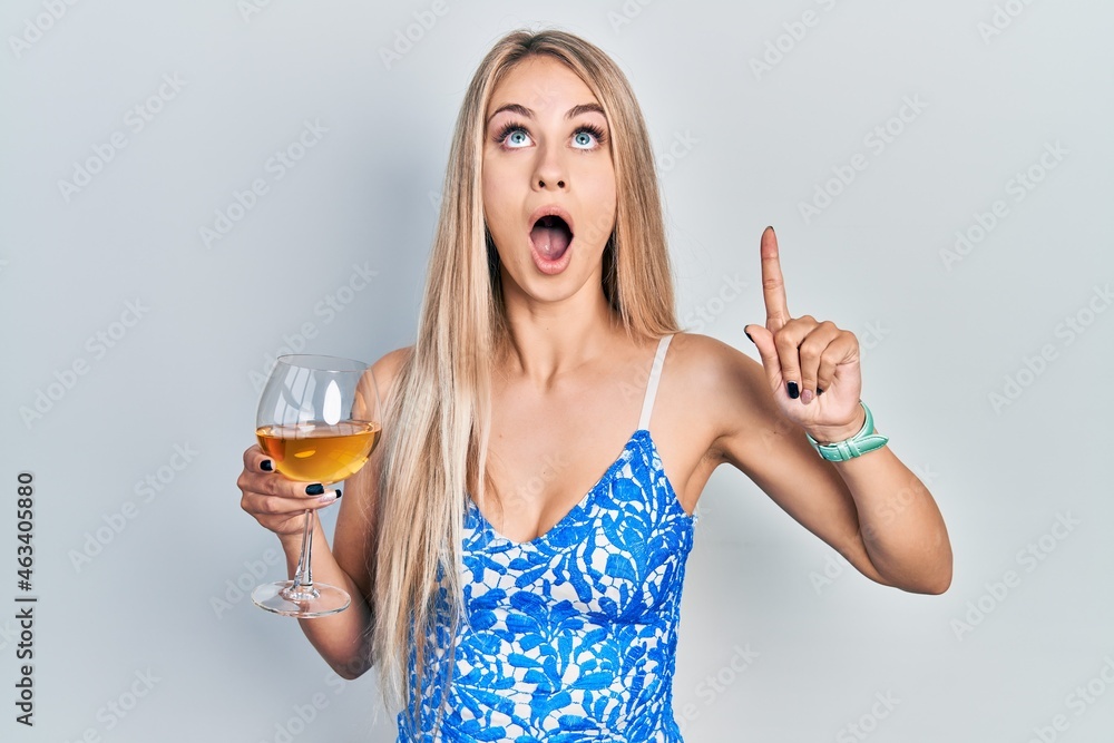 Young beautiful caucasian woman drinking a glass of white wine amazed and surprised looking up and pointing with fingers and raised arms. Young beautiful caucasian woman drinking a glass of white wine amazed and surprised looking up and pointing with fingers and raised arms.