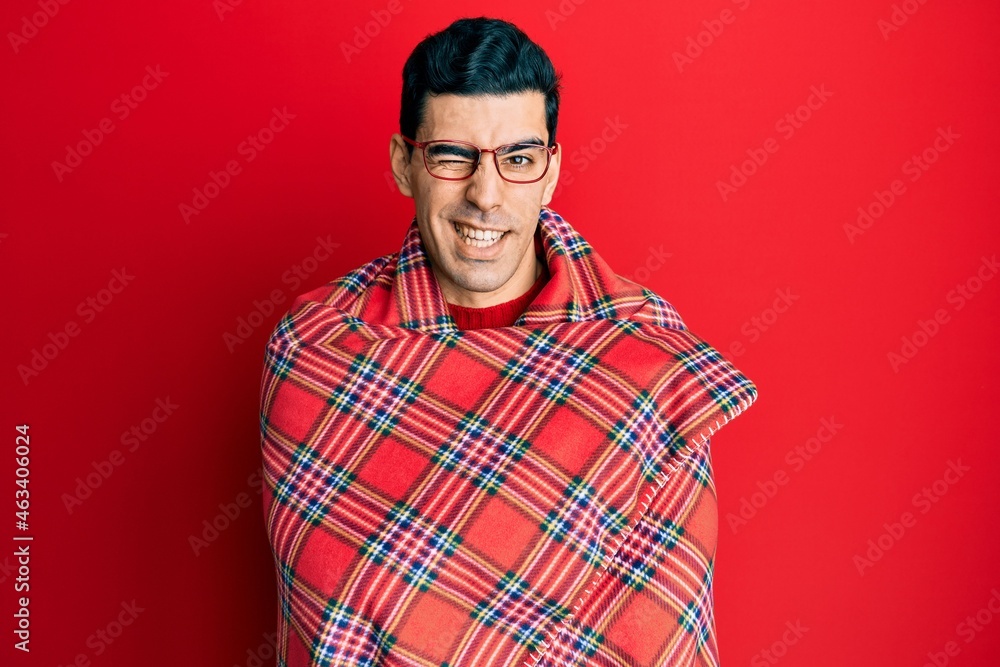 Handsome hispanic man wrapped in a red warm red blanket winking looking at the camera with sexy expression, cheerful and happy face. Handsome hispanic man wrapped in a red warm red blanket winking looking at the camera with sexy expression, cheerful and happy face.