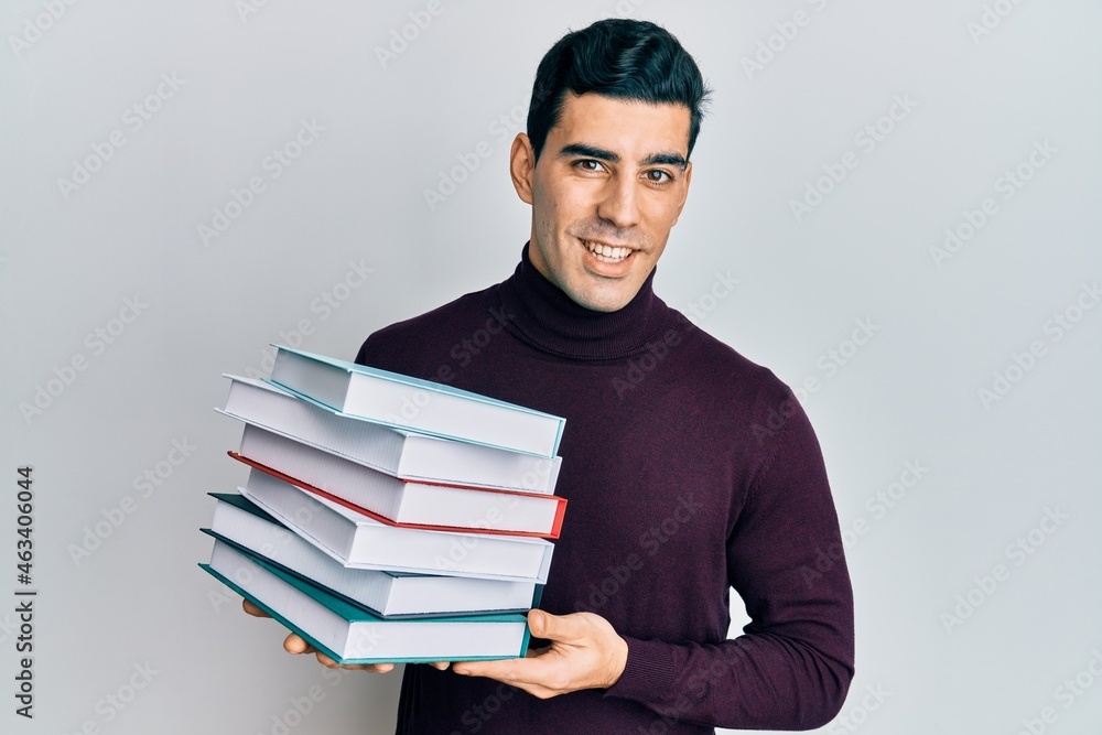 Handsome hispanic man holding a pile of books looking positive and happy standing and smiling with a confident smile showing teeth
