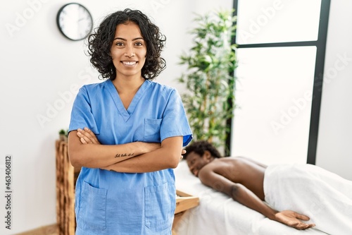 Young physiotherapist woman smiling happy standing with arms crossed gesture. at the clinic. African american man waiting to recive massage at the clinic.