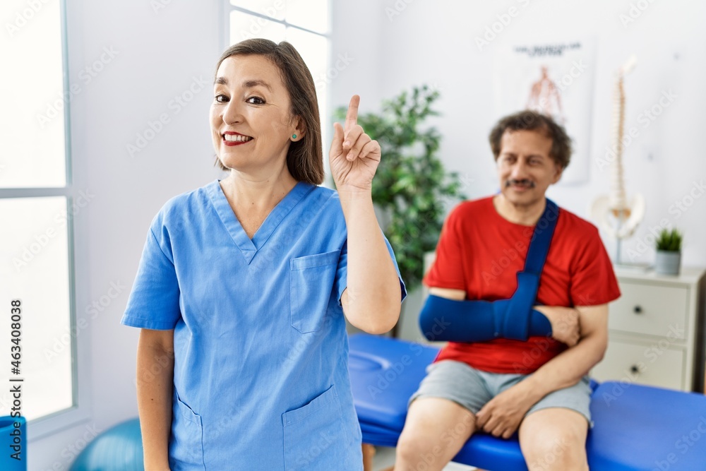 Fototapeta premium Middle age doctor woman with patient with arm injury at rehabilitation clinic smiling with an idea or question pointing finger up with happy face, number one