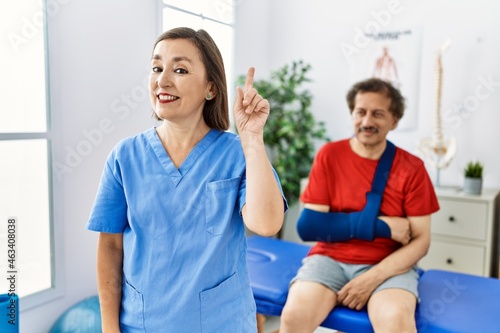 Middle age doctor woman with patient with arm injury at rehabilitation clinic smiling with an idea or question pointing finger up with happy face  number one