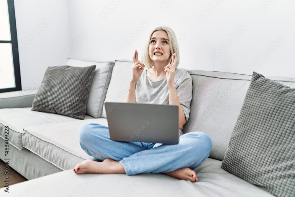 Young caucasian woman using laptop at home sitting on the sofa gesturing finger crossed smiling with hope and eyes closed. luck and superstitious concept. Young caucasian woman using laptop at home sitting on the sofa gesturing finger crossed smiling with hope and eyes closed. luck and superstitious concept.