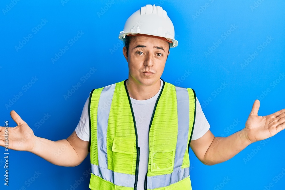 Handsome young man wearing safety helmet and reflective jacket clueless and confused expression with arms and hands raised. doubt concept. Handsome young man wearing safety helmet and reflective jacket clueless and confused expression with arms and hands raised. doubt concept.