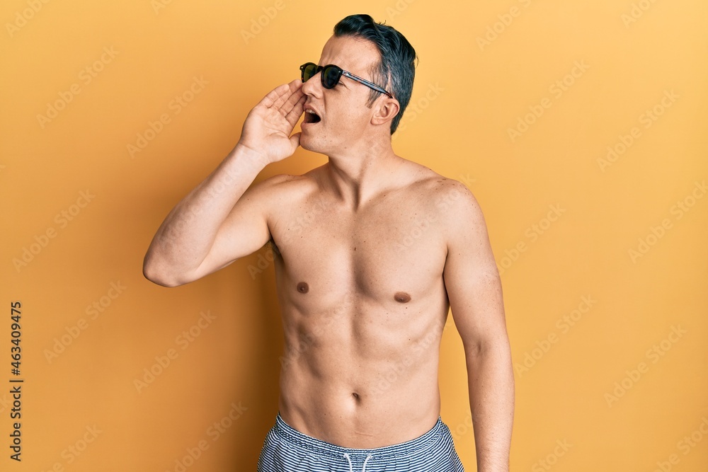 Handsome young man wearing swimsuit and sunglasses shouting and screaming loud to side with hand on mouth. communication concept.