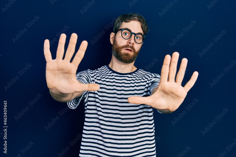 Naklejka premium Caucasian man with beard wearing striped t shirt and glasses afraid and terrified with fear expression stop gesture with hands, shouting in shock. panic concept.