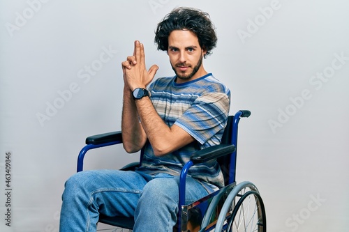 Handsome hispanic man sitting on wheelchair holding symbolic gun with hand gesture, playing killing shooting weapons, angry face