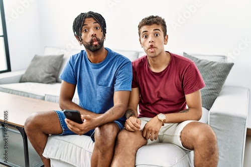 Young hispanic men using smartphone sitting on the sofa at home making fish face with lips, crazy and comical gesture. funny expression.