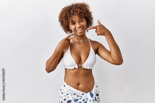 Young african american woman with curly hair wearing bikini smiling cheerful showing and pointing with fingers teeth and mouth. dental health concept.