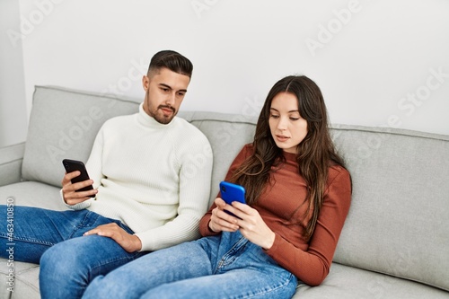 Young hispanic couple using smartphone sitting on the sofa at home.
