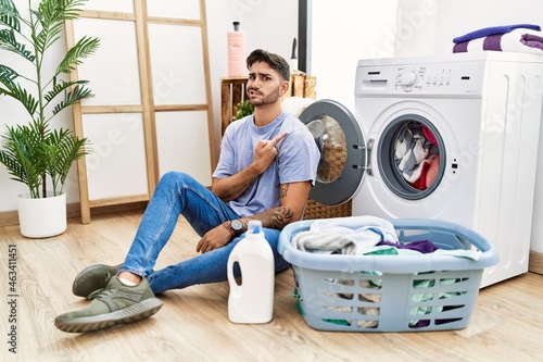 Young hispanic man putting dirty laundry into washing machine pointing aside worried and nervous with forefinger, concerned and surprised expression
