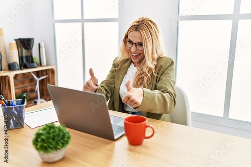 Beautiful blonde woman working at the office with laptop approving doing positive gesture with hand, thumbs up smiling and happy for success. winner gesture.