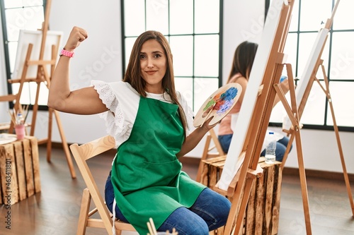 Young hispanic artist women painting on canvas at art studio strong person showing arm muscle, confident and proud of power