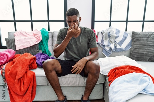 Young african american man sitting on the sofa with dirty laundry clothes smelling something stinky and disgusting, intolerable smell, holding breath with fingers on nose. bad smell