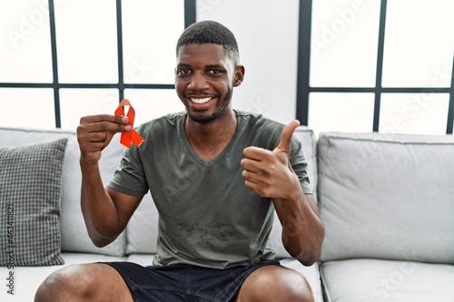 Young african american man holding support red ribbon sitting on the sofa smiling happy and positive, thumb up doing excellent and approval sign © Krakenimages.com