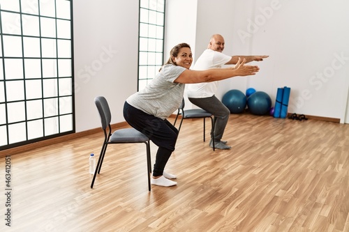 Middle age hispanic couple stretching using chair at sport center.