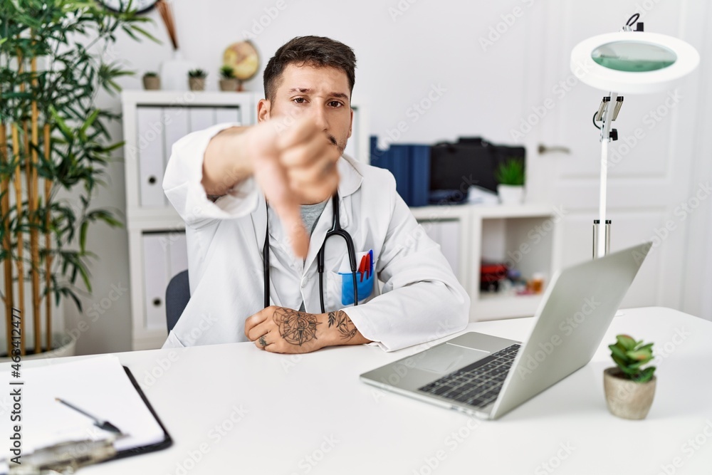 Young doctor working at the clinic using computer laptop looking unhappy and angry showing rejection and negative with thumbs down gesture. bad expression.