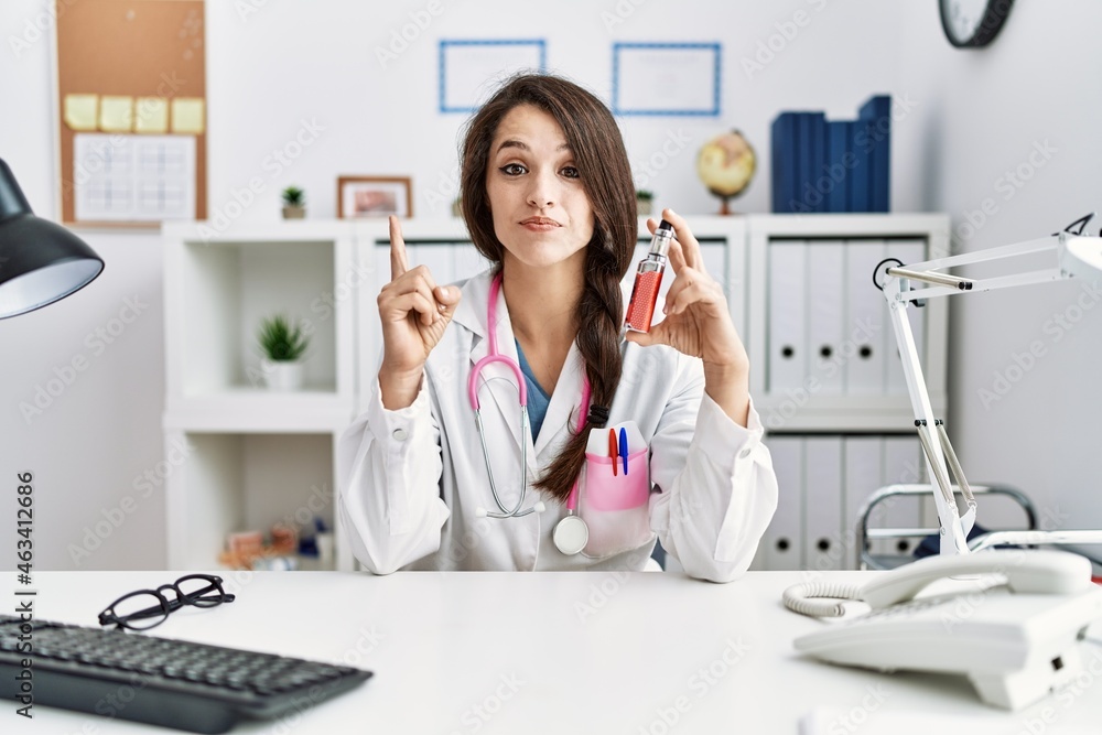 Young doctor woman holding electronic cigarette at medical clinic surprised with an idea or question pointing finger with happy face, number one Young doctor woman holding electronic cigarette at medical clinic surprised with an idea or question pointing finger with happy face, number one