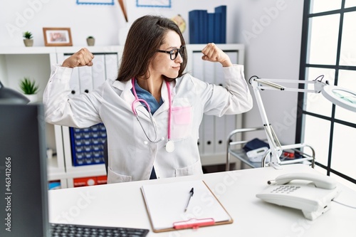Young doctor woman wearing doctor uniform and stethoscope at the clinic showing arms muscles smiling proud. fitness concept.